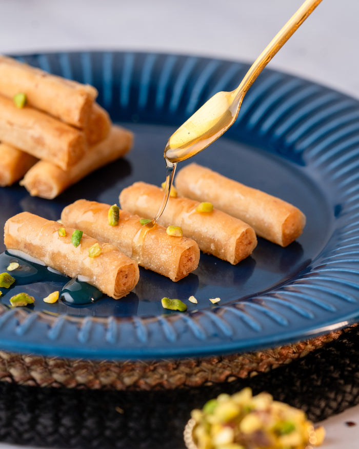 Lady finger baklava rolls on a blue plate with syrup poured from a gold spoon